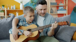 Adorable boy is playing the guitar under guidance of caring father learning at home together sitting on couch in cozy room. People and music concept.