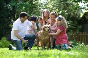 Keeping your furry pets healthy includes keeping them clean, as well. Together, this family was in the process of washing their Labrador retriever outside in the fresh air. With help from two neighbor girls, the mother, young daughter, and son, were all soaping down the dog’s coat, while the father was steadying the pet using a leash and his hand.