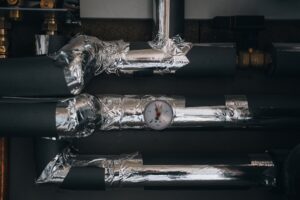 A network of insulated heating or plumbing pipes, wrapped in silver foil tape, features a central pressure gauge indicating system status. The image highlights industrial efficiency and technical precision in utility infrastructure.