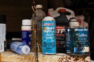 A Row of Assorted Cans and Bottles on a Rusty Shelf in a Garage - An assortment of cans and bottles, including a blue can with Quick Start in yellow letters, a black can marked ATF, and a blue and white can labeled Beautifone Tableau Peinture. The items are lined up on a rusty, wooden shelf, likely in a garage or workshop.