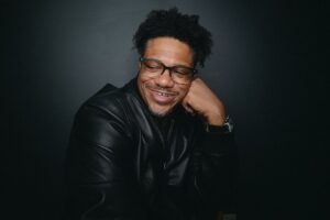 A black man with a confident smile, wearing glasses and a black leather jacket, rests his head on his hand against a dark background. The photo exudes a relaxed yet self-assured vibe, lifestyle image blending professionalism with warmth.