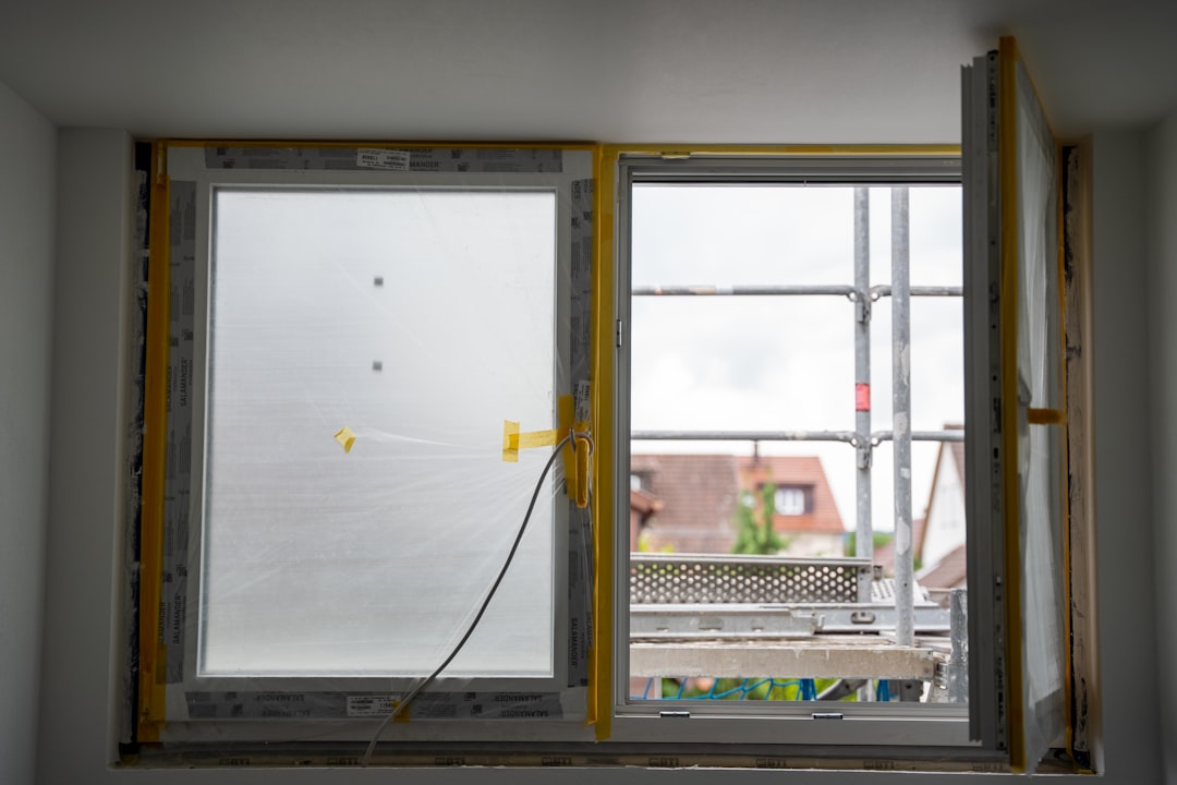 A close-up view of a partially installed window, with one pane covered in protective plastic and the other revealing a scaffolded exterior and rooftops beyond. This image captures a transitional phase in home renovation, where construction meets daylight.