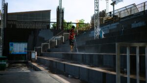 A worker uses a high-pressure power washer to clean concrete stadium seating at an outdoor venue. This wide-angle shot features a man in a red shirt performing maintenance on bleachers, highlighting industrial cleaning processes, facility management, and urban sanitation in a public space.